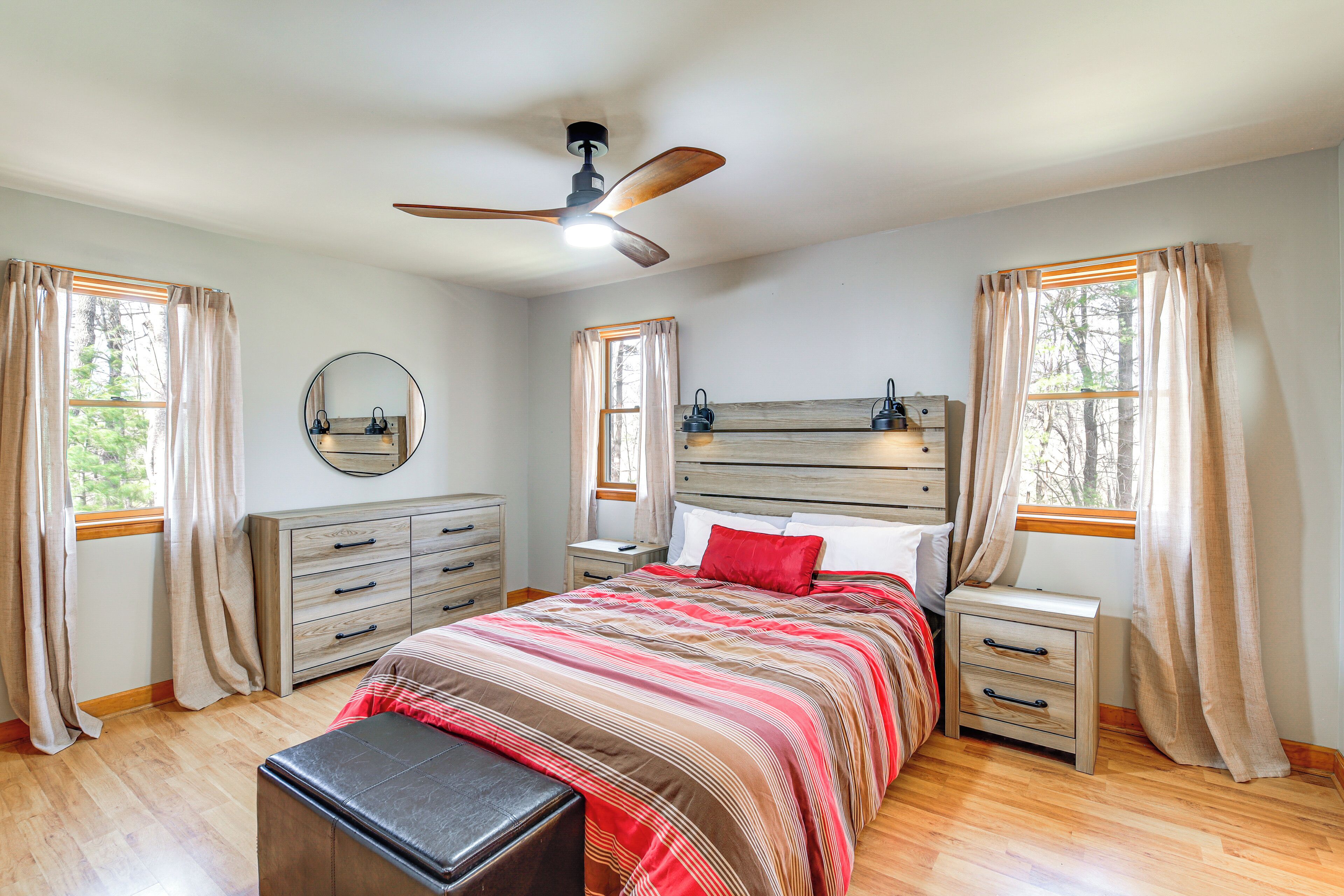 A bedroom with a bed, nightstands, a dresser, a ceiling fan, and large windows with curtains.