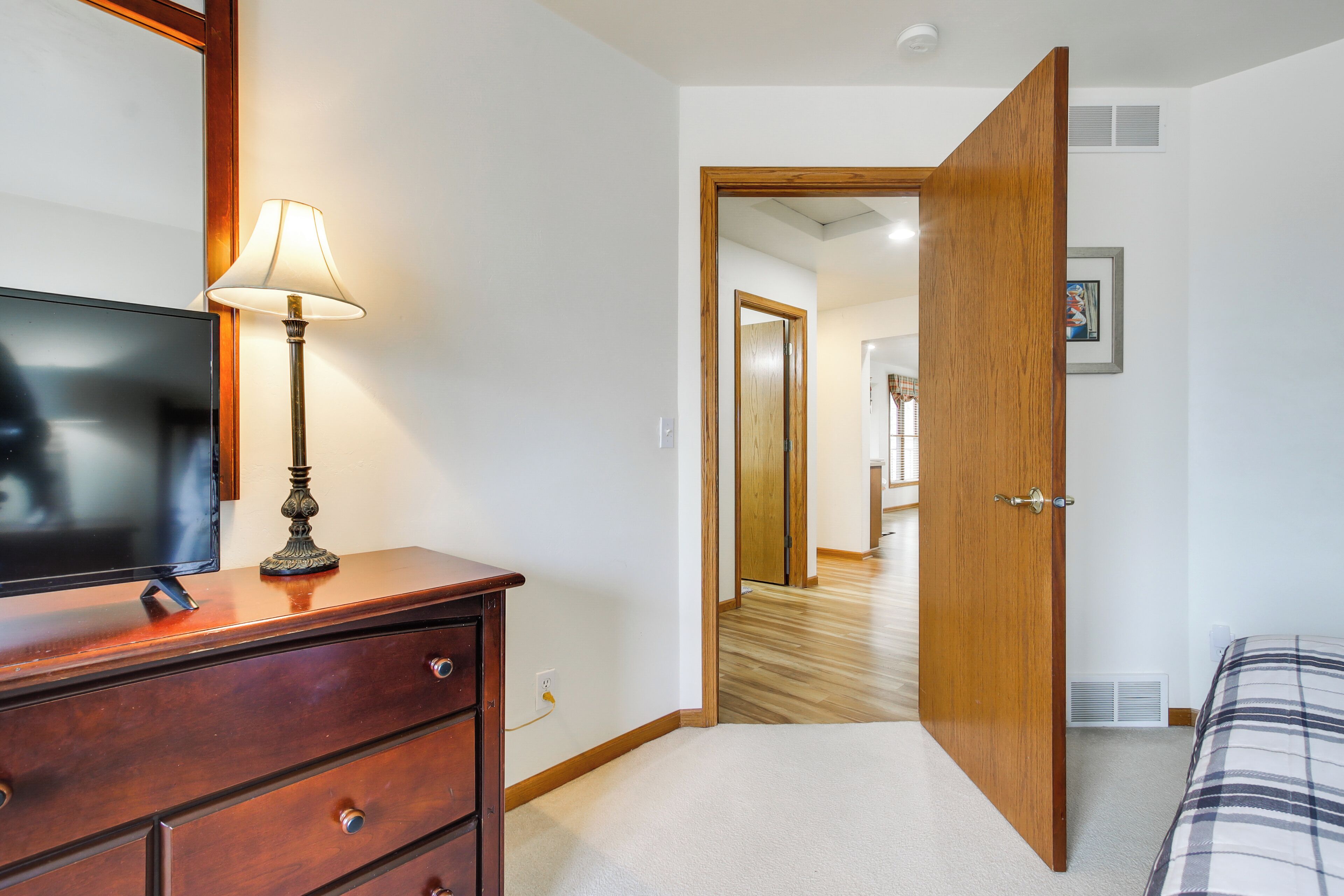 A bedroom with a wooden dresser, a lamp, a bed with a plaid bedspread, and a door leading to another room.