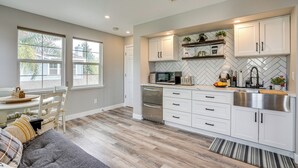A modern kitchen with white cabinets, a stainless steel sink, and a microwave. There is a dining area with a gray sofa and a small table.