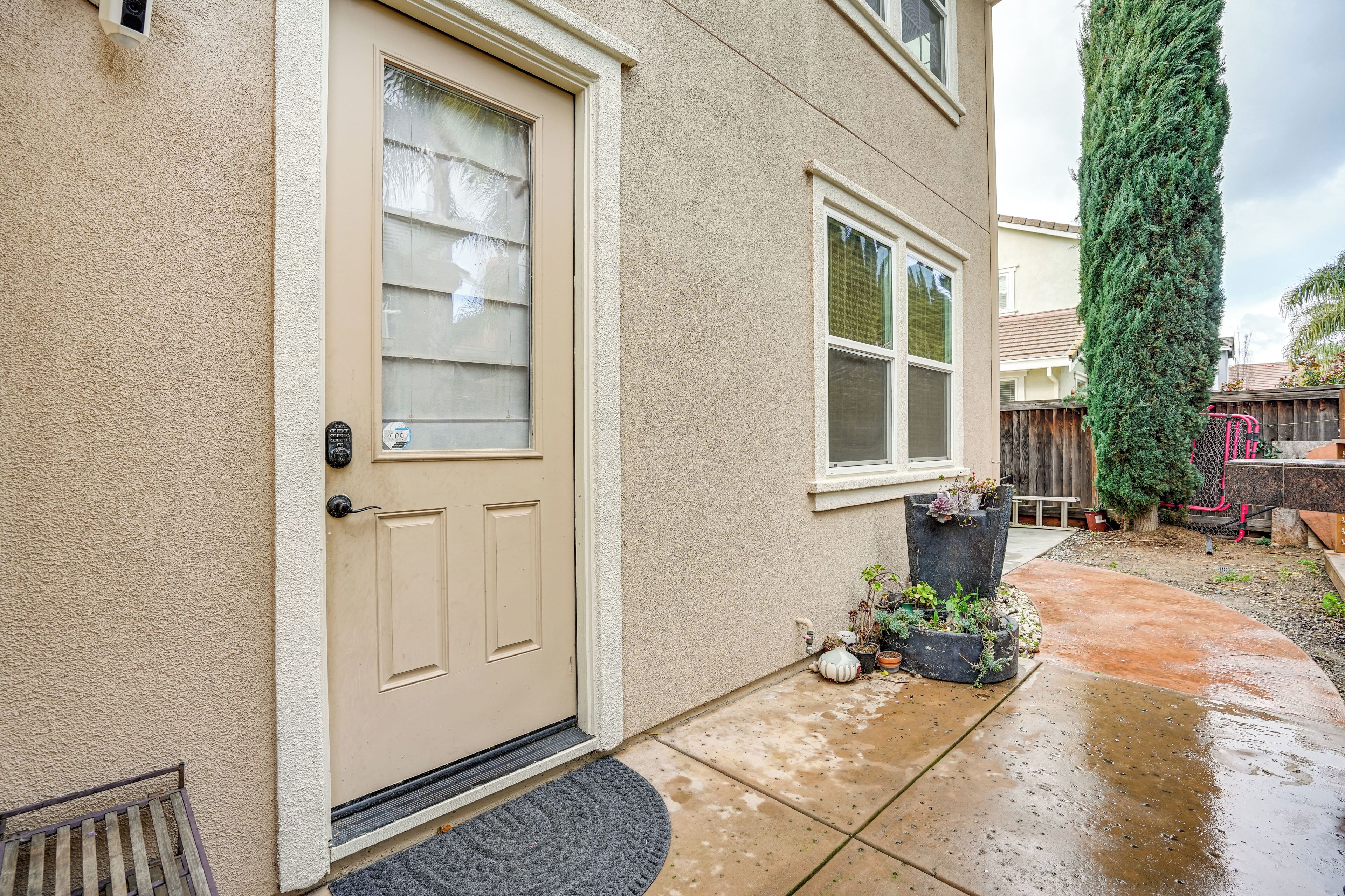A beige house with a front door, a window, and a small garden area with potted plants and a tall tree.