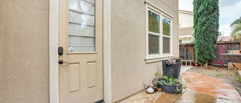 A beige house with a front door, a window, and a small garden area with potted plants and a tall tree.