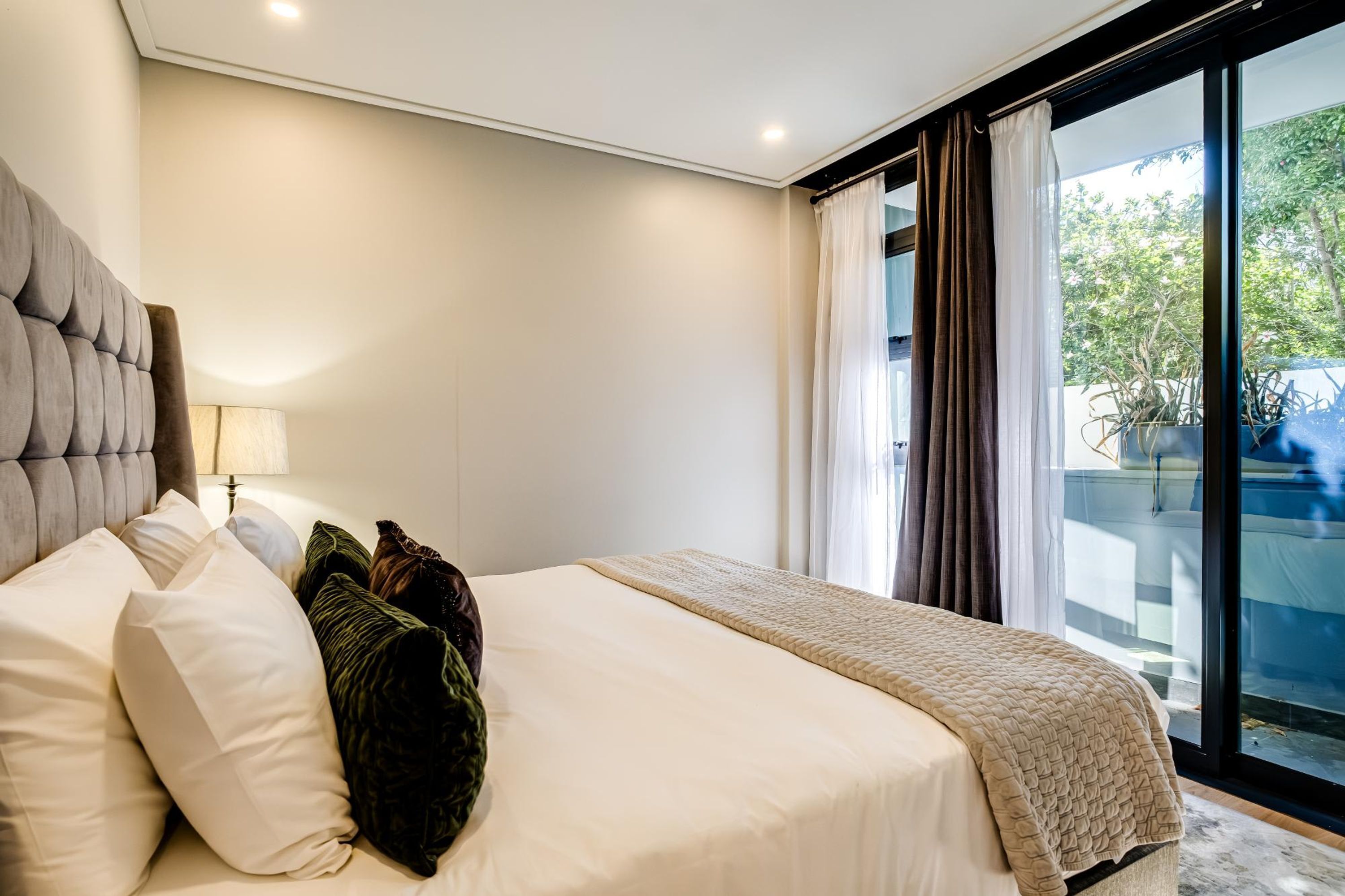 A bedroom with a large bed, a headboard, a bedside table with a lamp, and a view through a glass door to the outside.