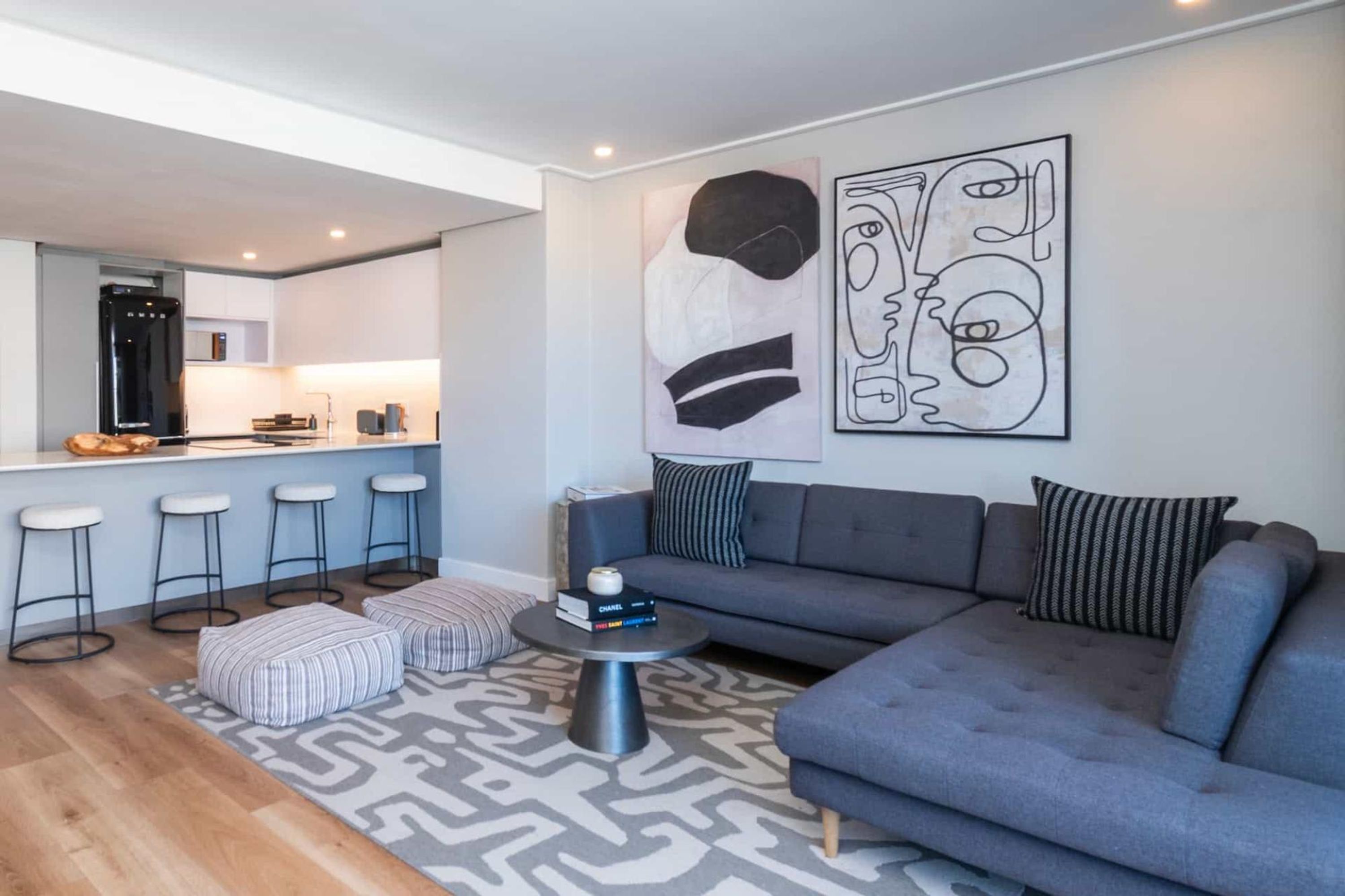 Una sala de estar moderna con un sofá gris, otomanas a rayas y una mesa de centro. Un cuadro abstracto grande decora la pared.