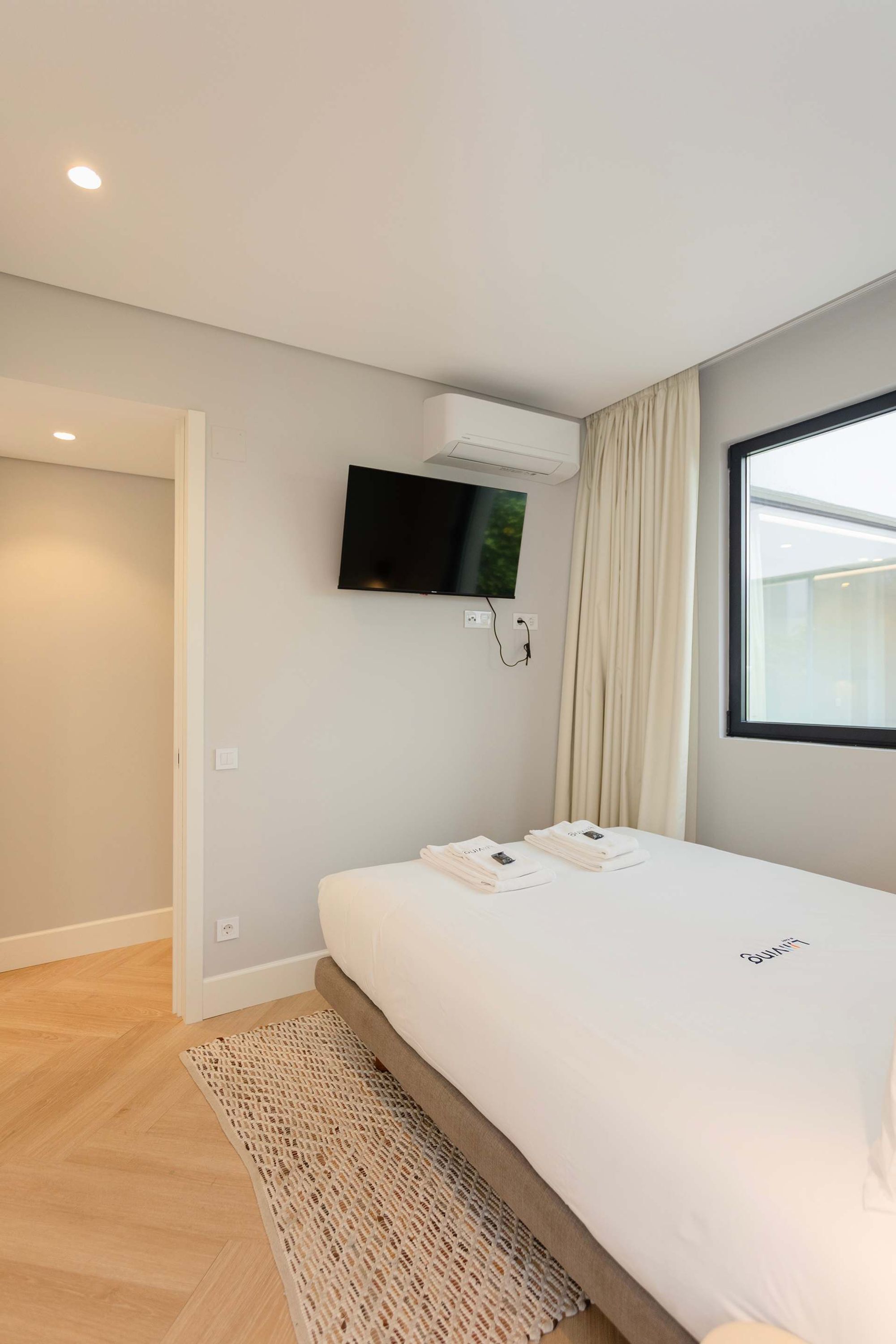 A modern hotel room with a large bed, a flat-screen TV mounted on the wall, and a window with curtains.