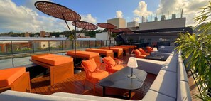 Terraza en la azotea con asientos naranjas, mesas y sombrillas, con vistas a un paisaje urbano.