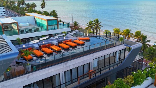 Een modern hotel met een zwembad op het dak, voorzien van oranje ligbedden en glazen hekwerk met uitzicht op het strand.
