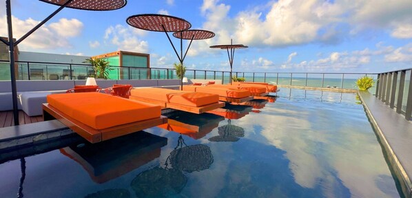 Una piscina infinita con tumbonas y sombrillas anaranjadas, rodeada de barandillas de cristal y una vista despejada del océano.