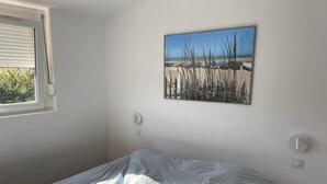 Een slaapkamer met een bed, een raam, een wandlamp en een schilderij van een strandgezicht.