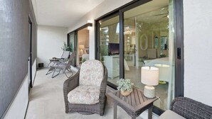 Un balcon moderne avec des meubles en osier, une table avec une lampe et une porte vitrée donnant sur un espace intérieur agrémenté d’un lustre.