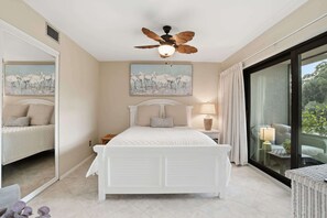 A bedroom with a bed, a ceiling fan, a nightstand with a lamp, and a large window with a view of the outdoors.