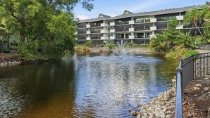 Un hôtel moderne avec une fontaine à l’avant, entouré d’une végétation luxuriante et sous un ciel dégagé.