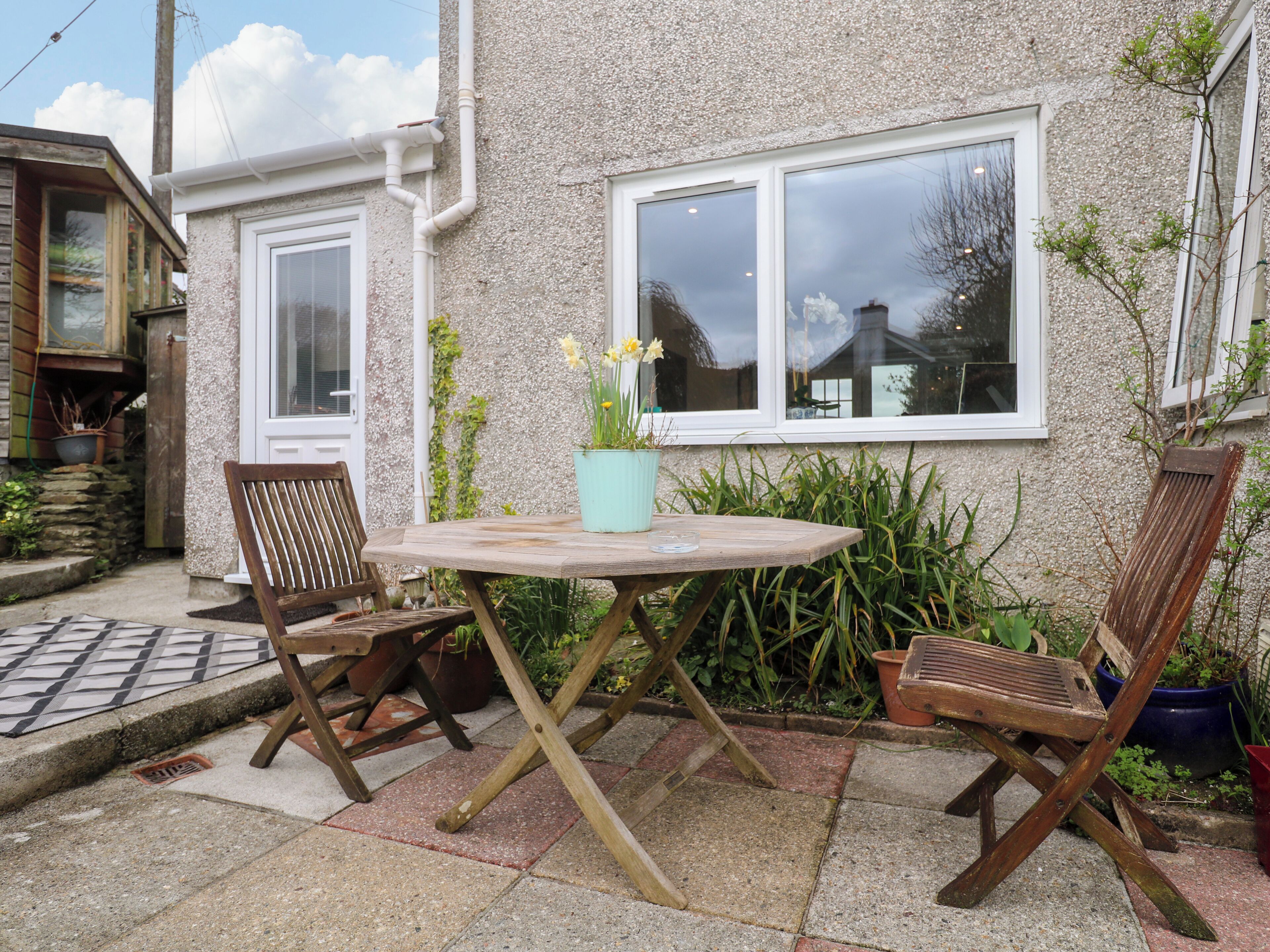 Un espace extérieur aménagé avec une table en bois et deux chaises, un pot de fleurs contenant des fleurs jaunes, et une maison avec une façade en pierre et une fenêtre.