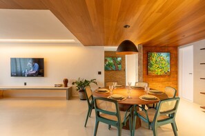 A modern dining area with a wooden ceiling, a large TV mounted on the wall, a dining table set with plates and glasses, and a potted plant. - Urban - Ed. Luciene (Recife)
