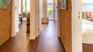 A hallway with wooden flooring, white walls, and artwork. There is a bed with a headboard visible on the right. - Urban - Ed. Luciene (Recife)