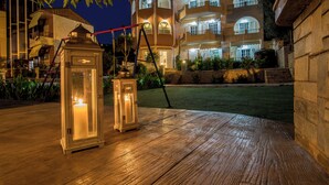 Un espace extérieur bien éclairé, doté d’une terrasse en bois, de deux lanternes illuminées, et, en arrière-plan, d’un grand bâtiment avec de nombreux balcons et fenêtres.