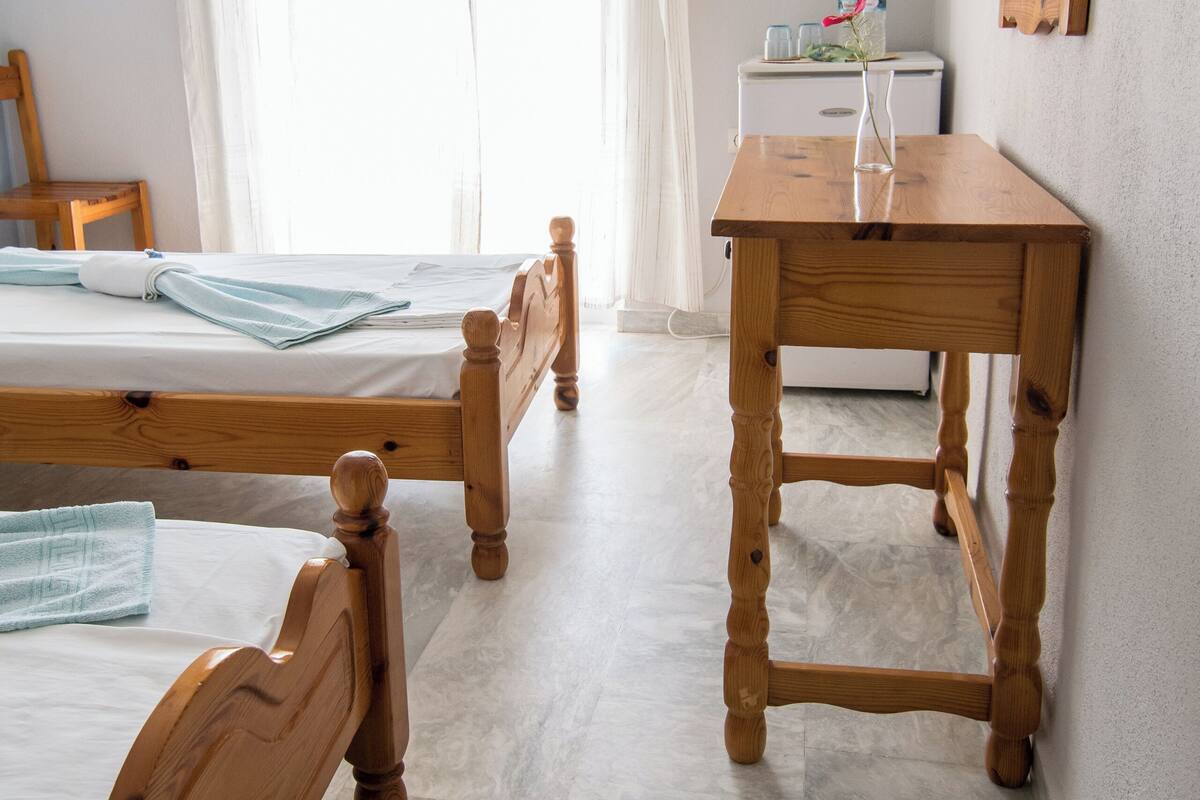 A room with two wooden beds, a wooden table, a TV mounted on the wall, and a window with curtains.