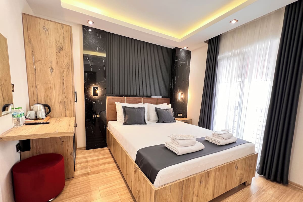 A modern hotel room with a wooden bed, a desk, a red ottoman, and a view of the city through large windows.
