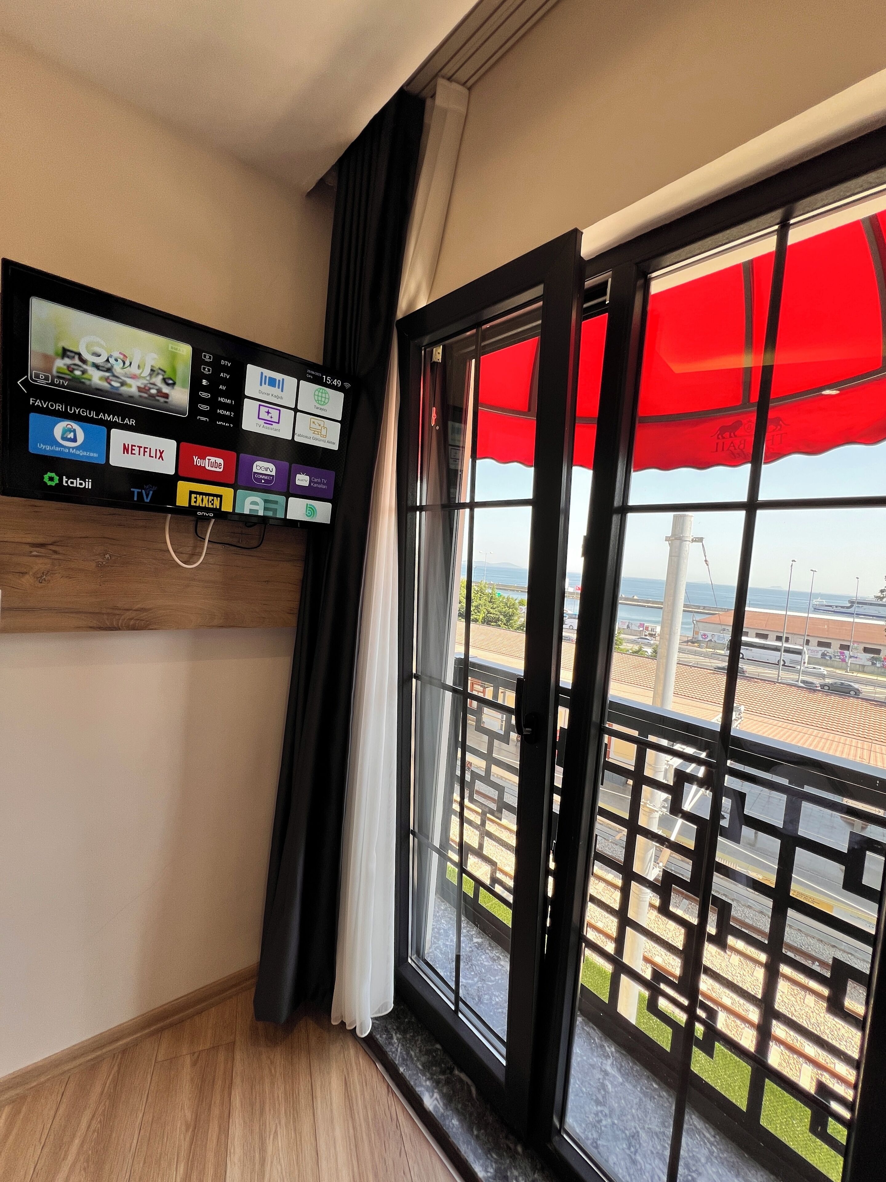 A modern hotel room with a large window, a TV mounted on the wall displaying various channels, and a balcony with a red awning.