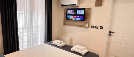 A modern hotel room with a large bed, a mounted TV displaying streaming services, and an air conditioning unit.