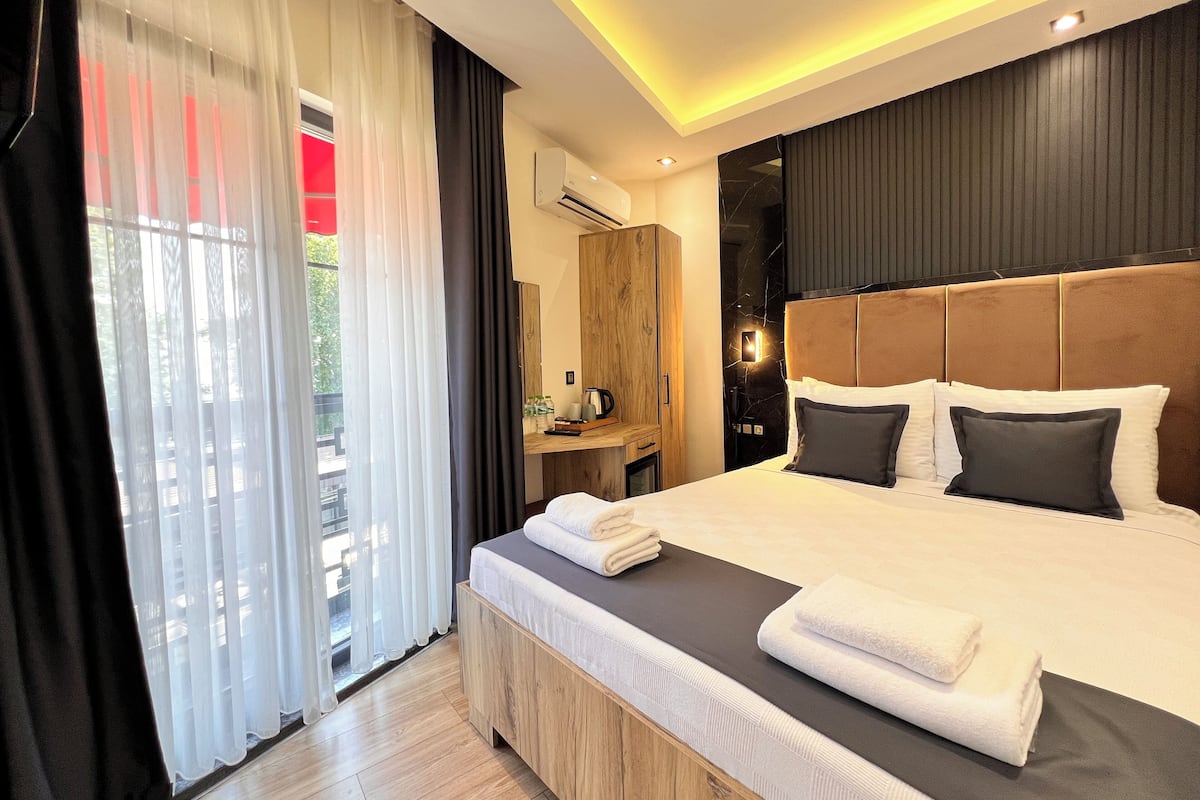 A modern hotel room with a large bed, a wooden bedside table, a mirror, and a view of the outdoors through sheer curtains.
