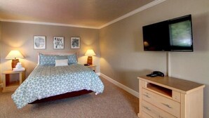 A bedroom with a bed, two bedside lamps, a flat-screen TV mounted on the wall, a wooden dresser, and a framed picture on the wall.