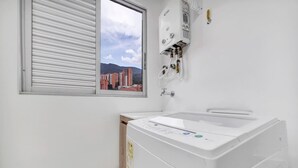 Une buanderie équipée d’un lave-linge à chargement frontal, d’une fenêtre avec des stores et d’un chauffe-eau fixé au mur.