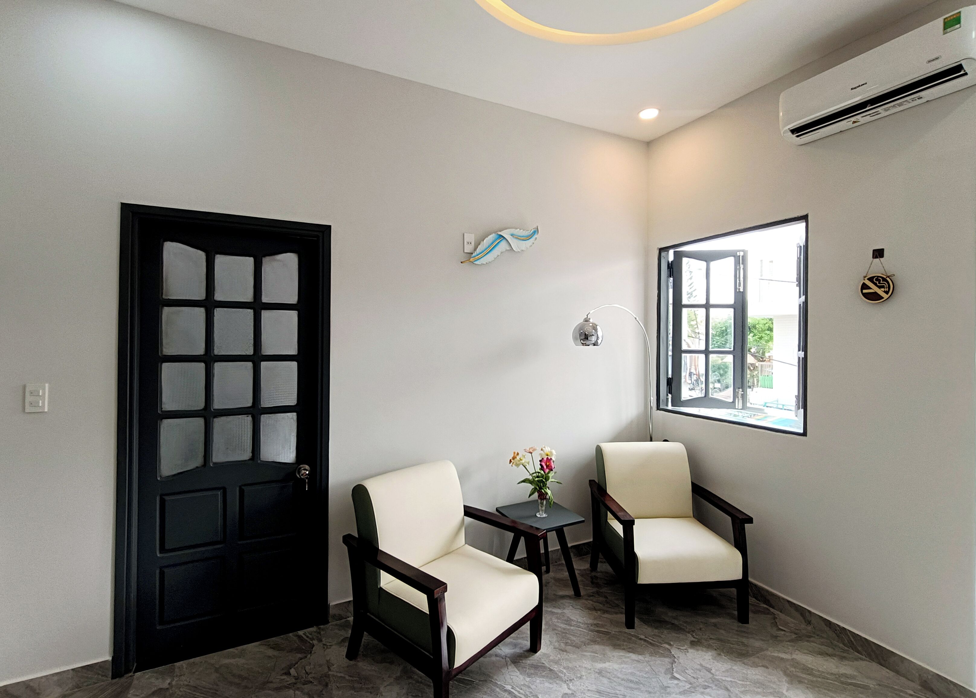 Une pièce avec une porte noire, deux chaises, une petite table sur laquelle se trouve un vase, une lampe fixée au mur et une fenêtre donnant sur un paysage.