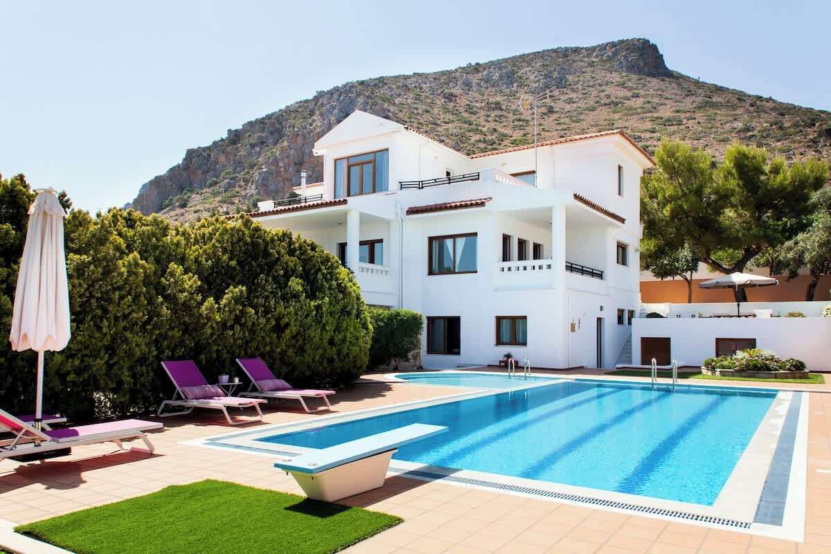 A two-story building with a swimming pool, lounge chairs, and a mountain backdrop.