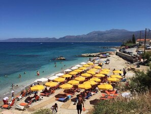 A beach with numerous yellow umbrellas, people swimming, and a clear view of the sea and mountains in the background. - Artemis Spacious Luxury Villa with XL Private Pool (Hersonissos)