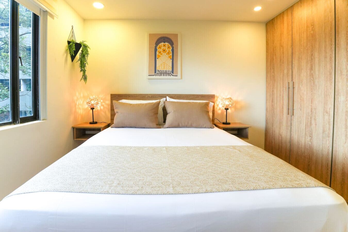 A large bed with a patterned bedspread, two bedside tables with lamps, a window with a view, and a wooden wardrobe.