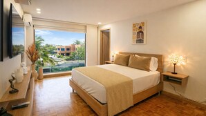 Un dormitorio moderno con una cama grande, suelo de madera, una mesita de noche con lámpara, un televisor montado en la pared y una ventana con vistas a edificios y zonas verdes.