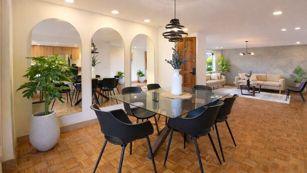 Un área de comedor moderna con una mesa de vidrio y sillas negras, rodeada de piso de madera y plantas en macetas.