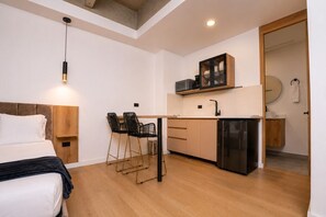 A compact hotel room with a bed, a small kitchenette, a dining area with chairs, and a wooden floor.
