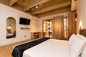 A modern hotel room with a large bed, a flat-screen TV mounted on the wall, a desk with a chair, and a window with curtains.