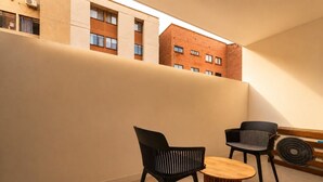 A modern interior with a round wooden table and two black chairs, a large window overlooking a brick building, and a wall-mounted fan.