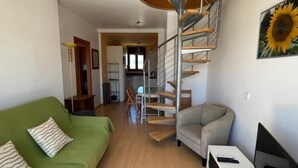 A modern living room with a green sofa, a grey armchair, a white TV stand, and a spiral staircase. - Beautiful duplex with sea view AV-72 (ROSES)