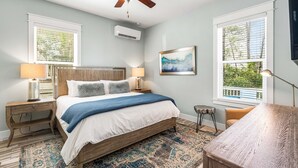 Un dormitorio con cama, dos mesitas de noche, ventilador de techo, equipo de aire acondicionado y una ventana grande con persianas.