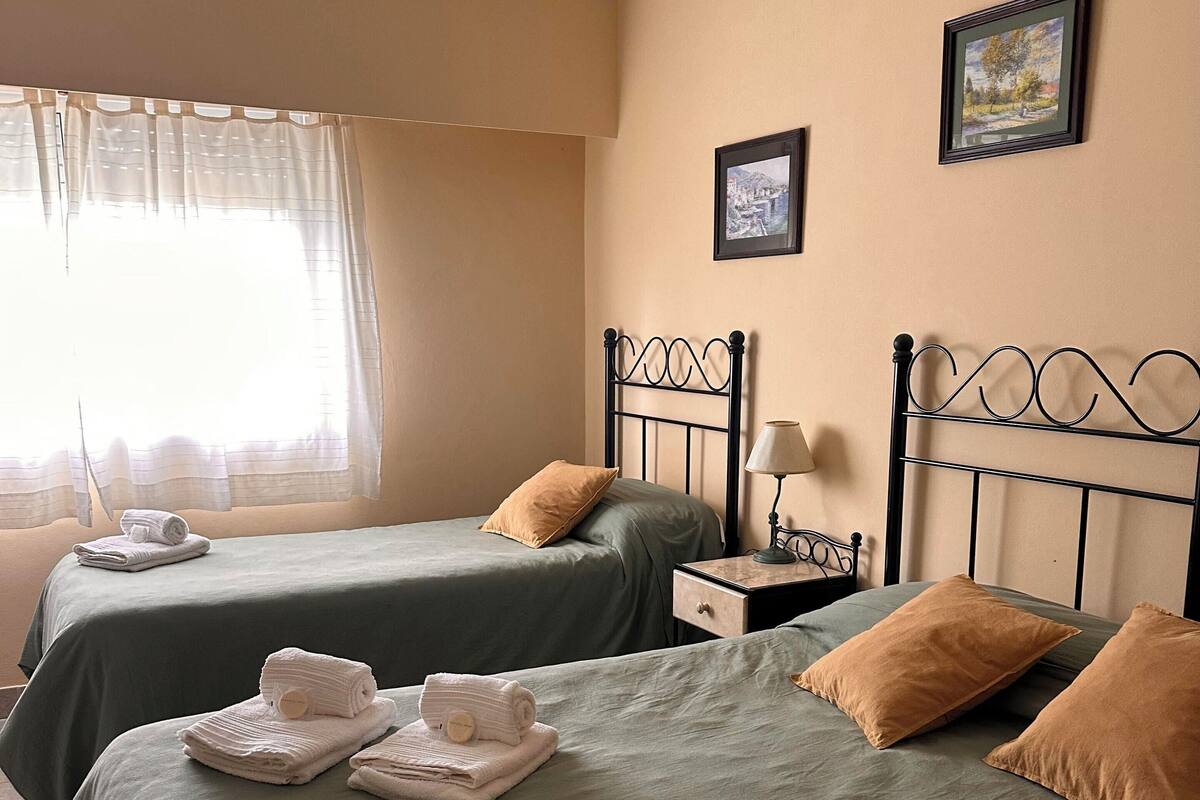 A hotel room with two beds, each with green bedding and a brown pillow. There is a nightstand with a lamp and a framed picture on the wall.
