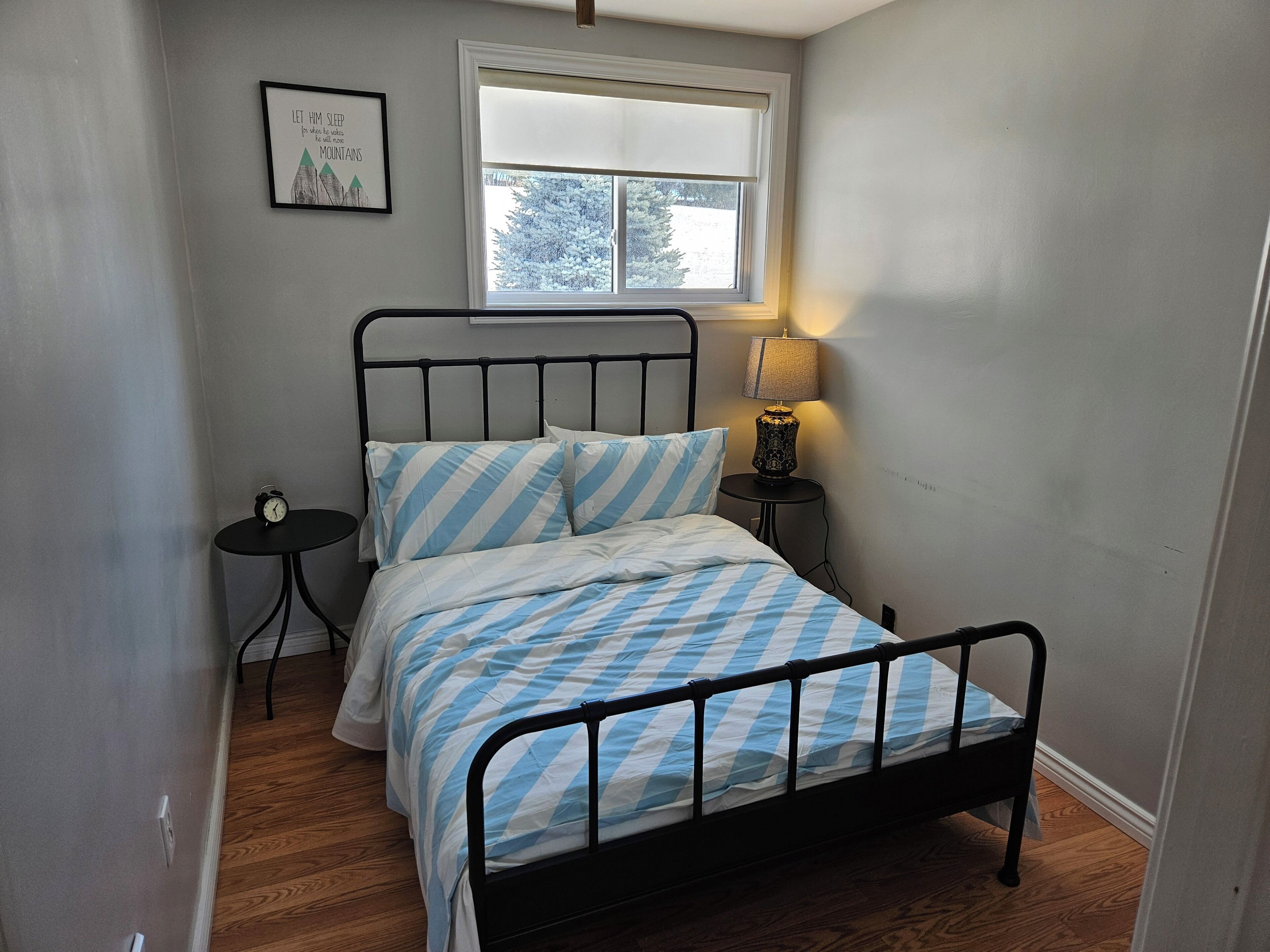 Une petite chambre à coucher, bien rangée, avec un lit, une table de chevet, une lampe et une citation encadrée accrochée au mur.