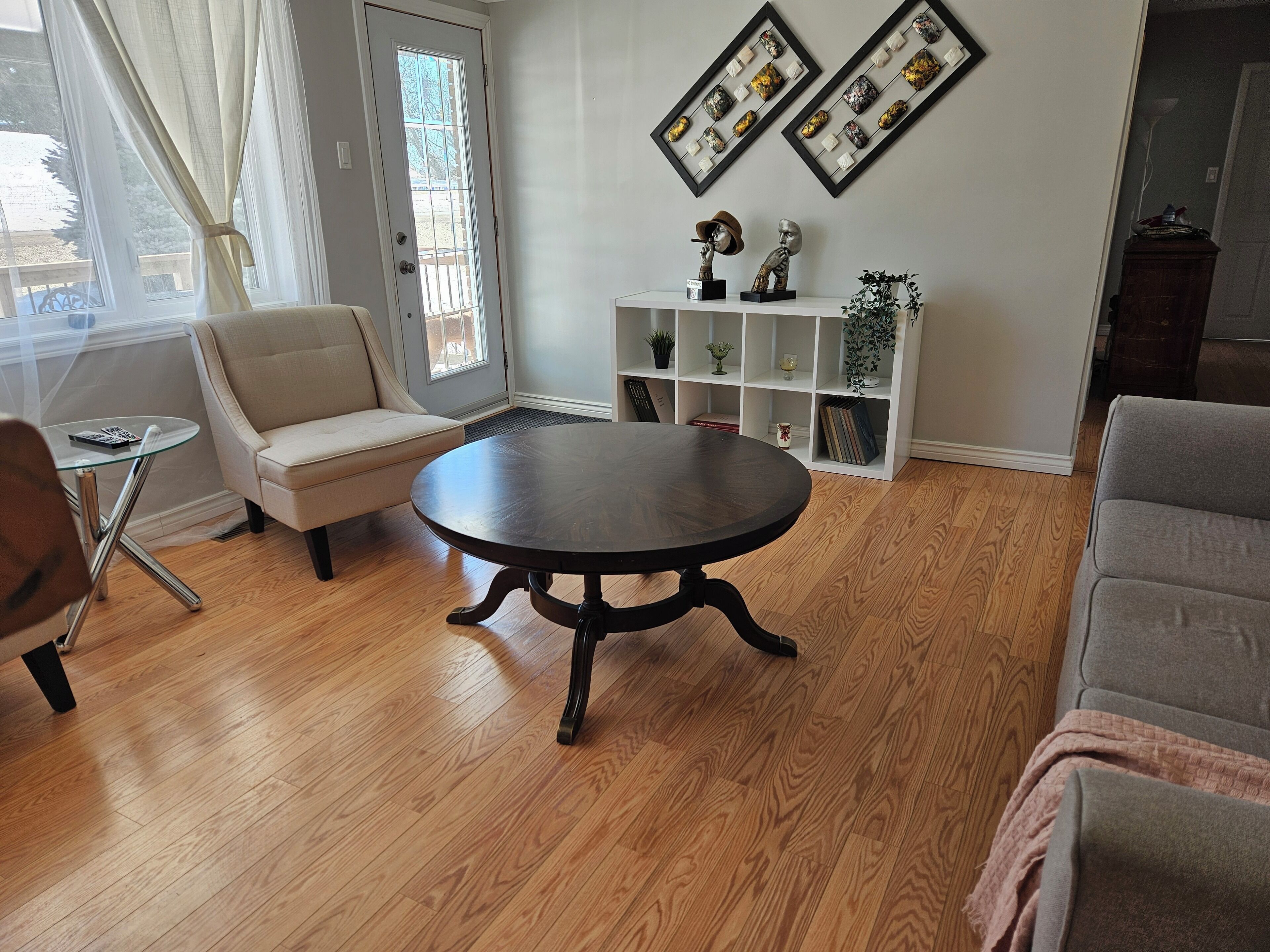 A living room with a round table, a beige sofa, a glass coffee table, and a white shelving unit with decorative items.