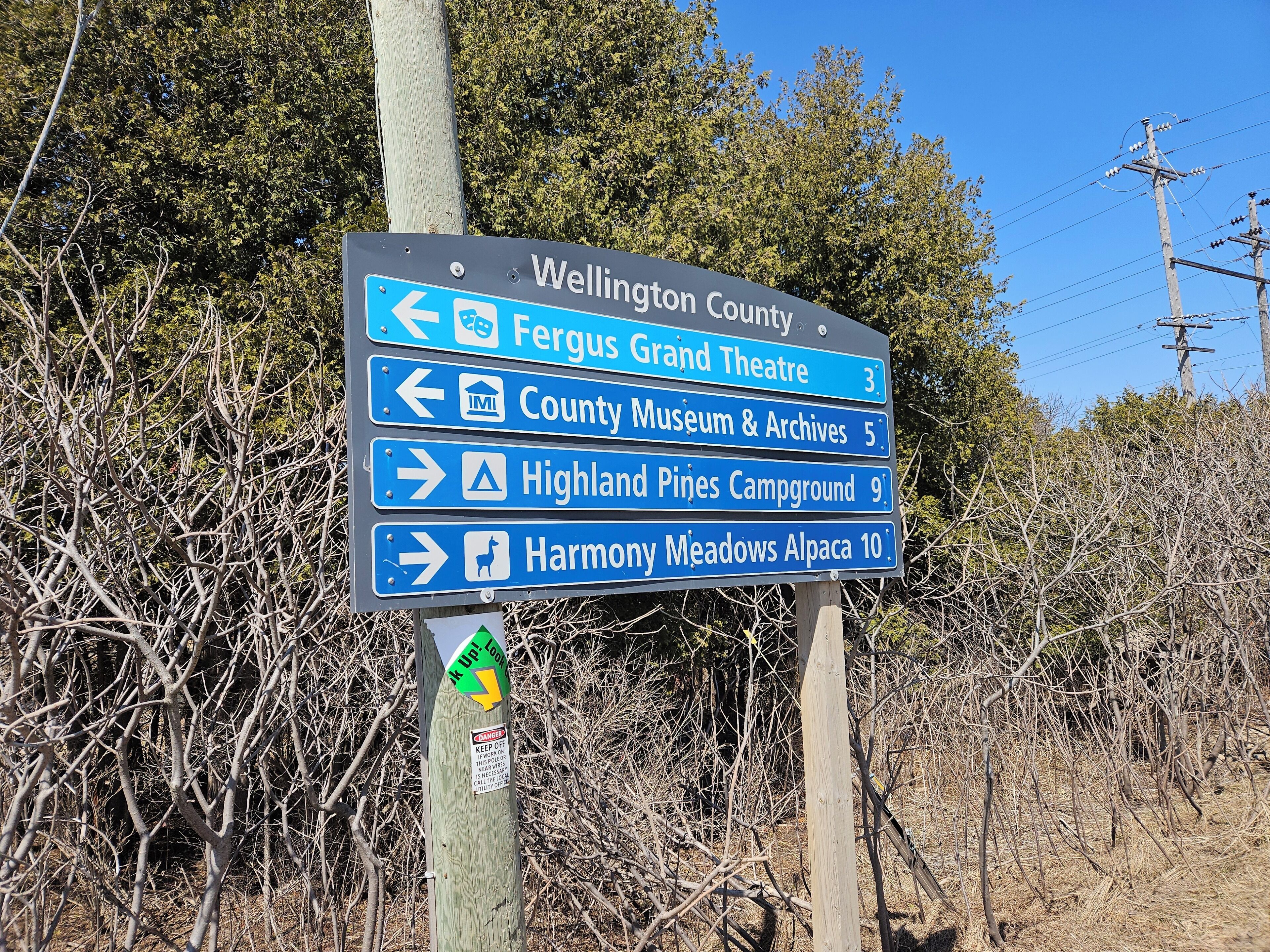 A directional signpost with Wellington County, Fergus Grand Theatre, County Museum & Archives, Highland Pines Campground, and Harmony Meadows Alpaca.