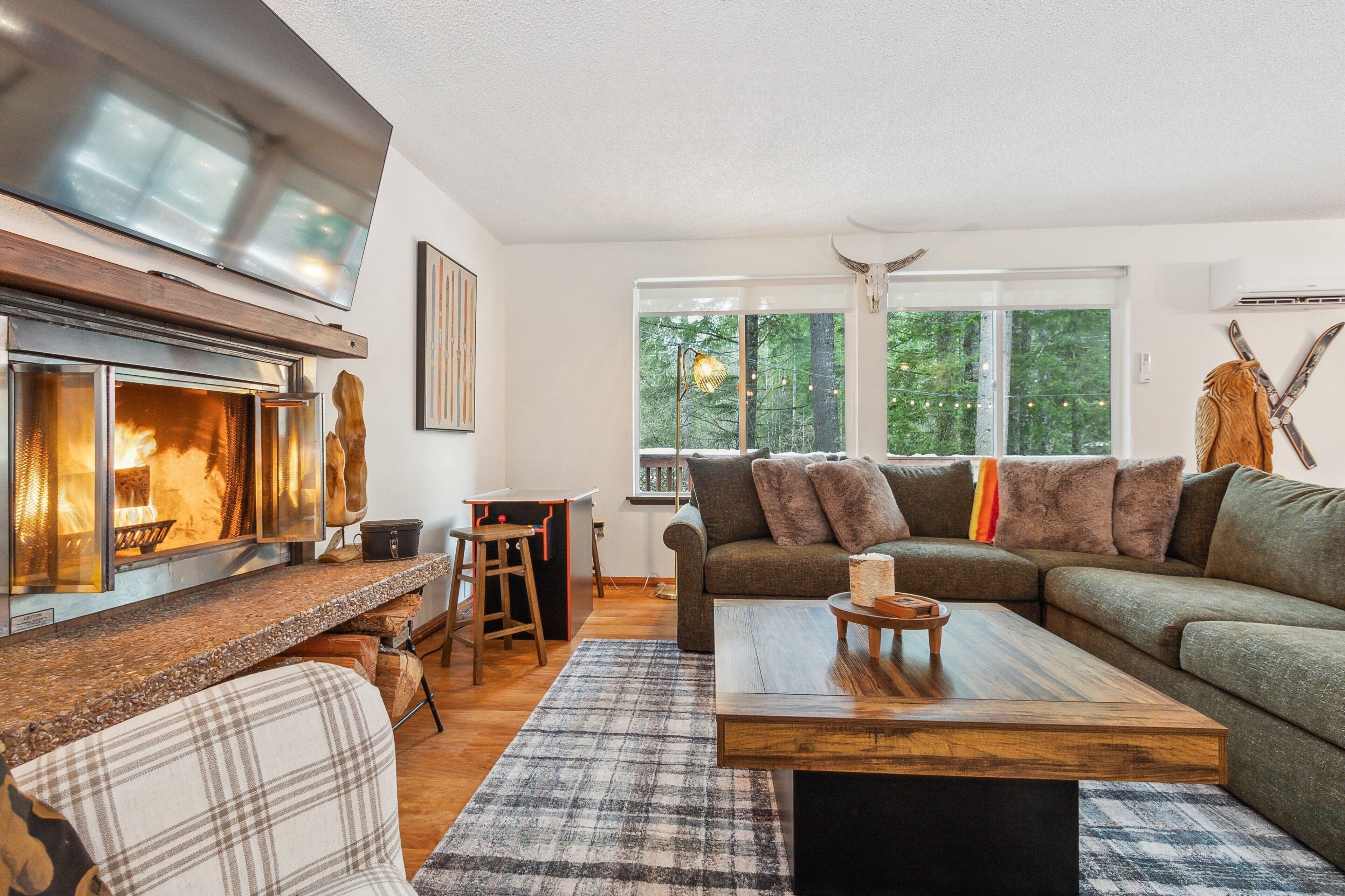 A cozy living room with a fireplace, a large window with a forest view, a sofa, and a coffee table.