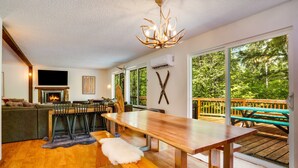 A modern interior with a wooden dining table, benches, a fireplace, and large windows overlooking a deck.