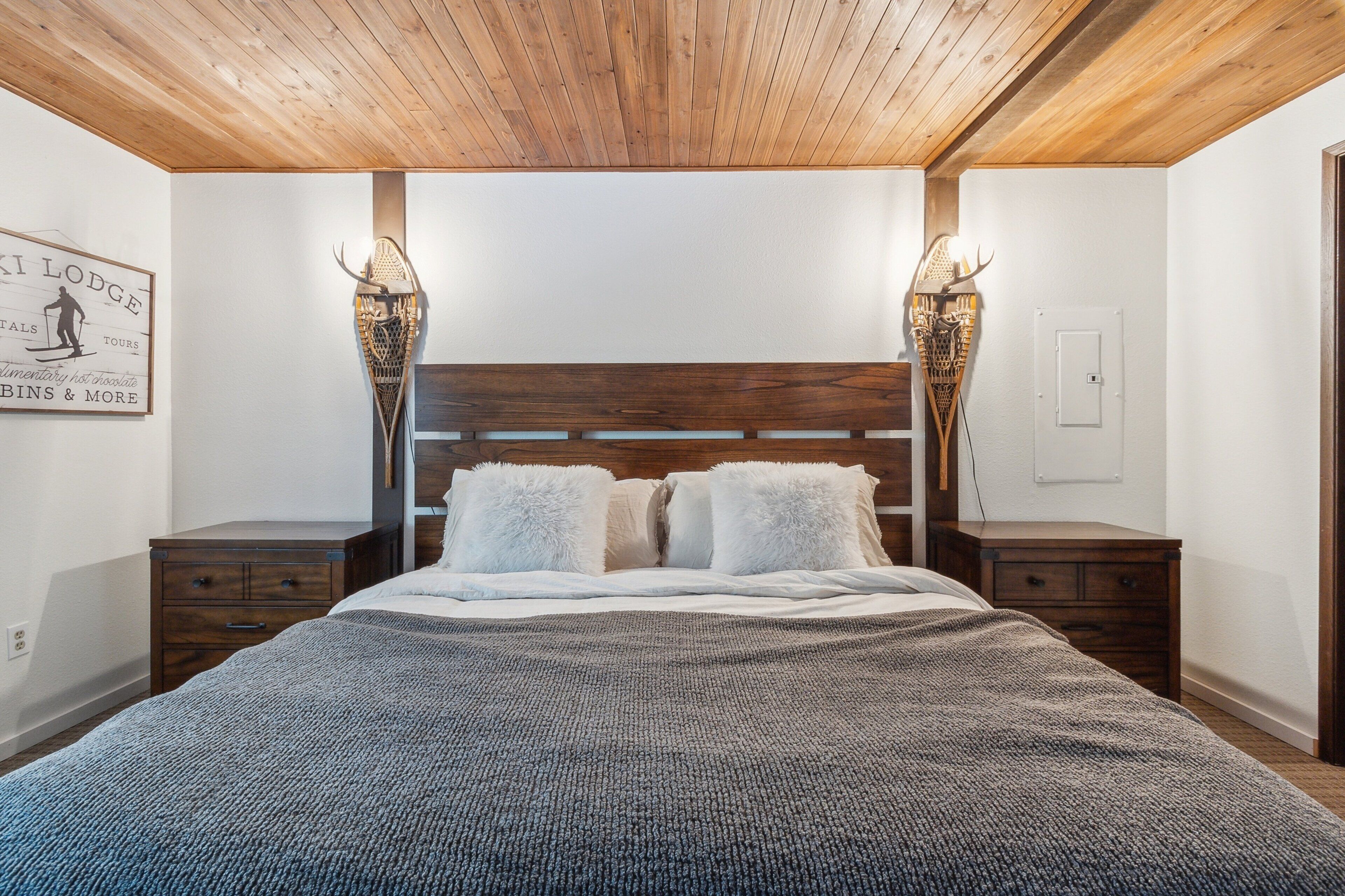 A bedroom with a wooden ceiling, a large bed with white bedding, two nightstands, and wall-mounted lamps.