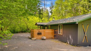 A cabin with a wooden deck, a gravel driveway, and a forested backdrop.