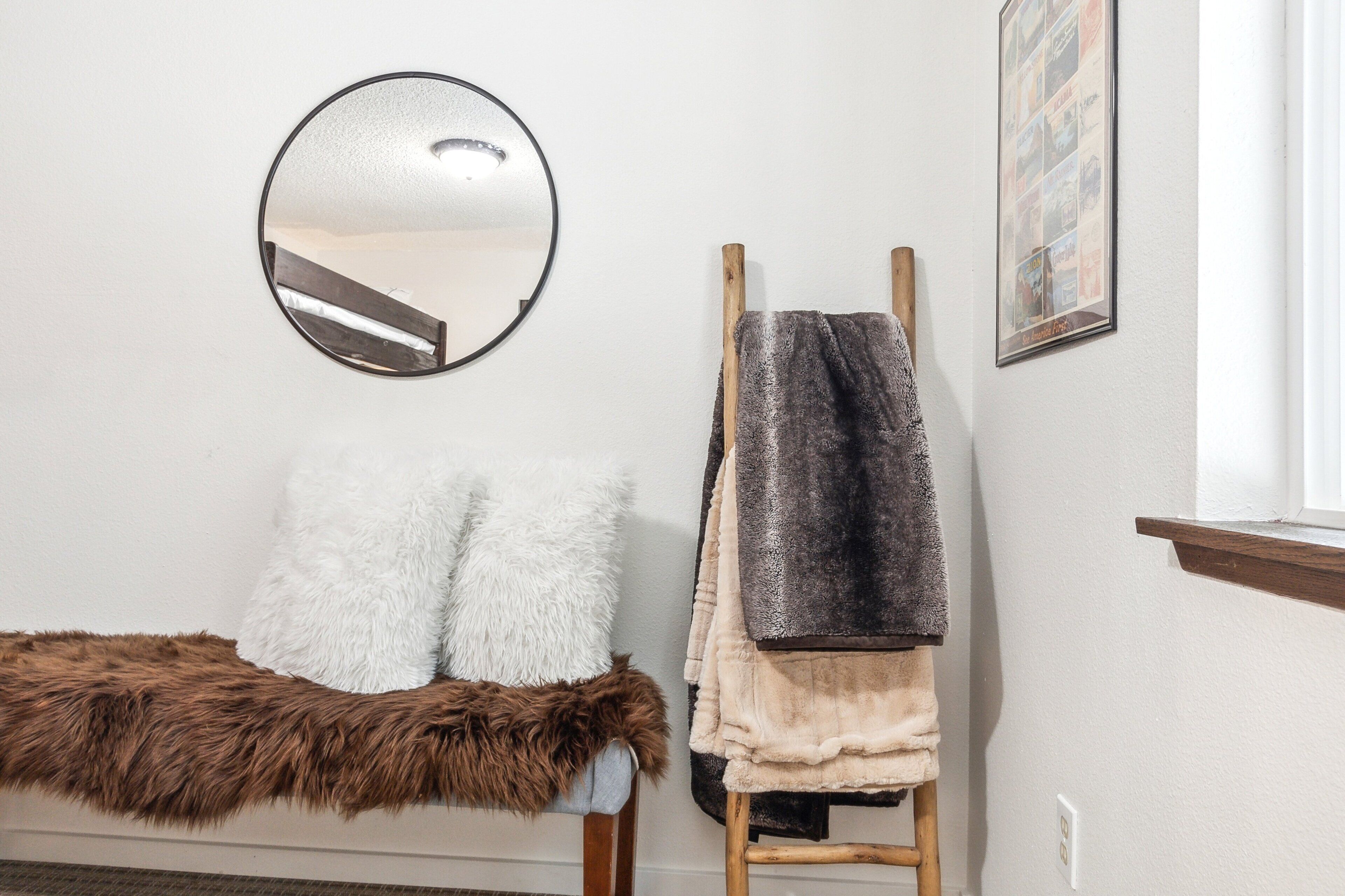 A room with a round mirror, a bench with fluffy pillows, a wooden stool with towels, and a framed picture on the wall.