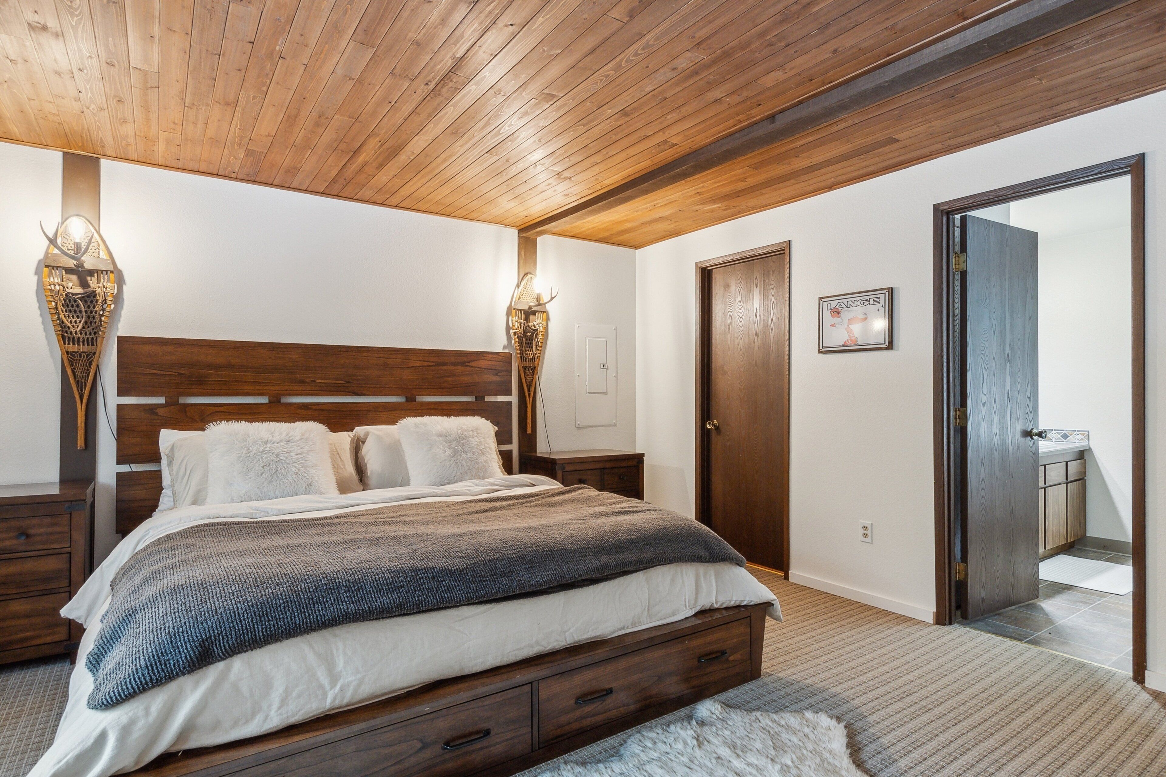 Un dormitorio con techo de madera, una cama grande con cabecera, dos lámparas fijadas a la pared, una puerta y un pequeño cuadro enmarcado en la pared.