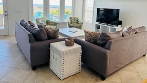 A living room with two sofas, a coffee table, a TV, and a large window with a view. - Fins / 1295 Ocean Blvd West 10 Bedroom Home (Supply)