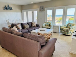 Uma sala de estar com sofá seccional, mesa de centro, duas poltronas, mesa de jantar com cadeiras e janelas amplas com vista para a praia.