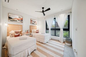A bedroom with two beds, a ceiling fan, artwork on the walls, and a window with curtains.